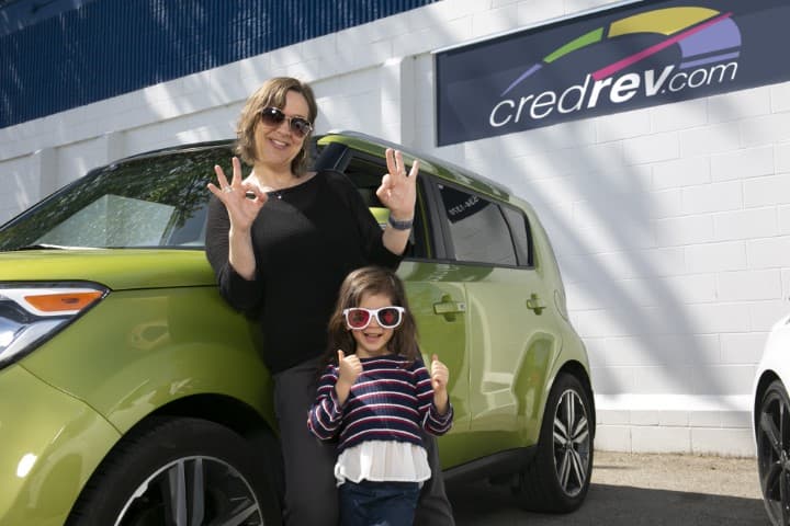 A woman and a child standing beside a green hatchback outside a CredRev-branded building